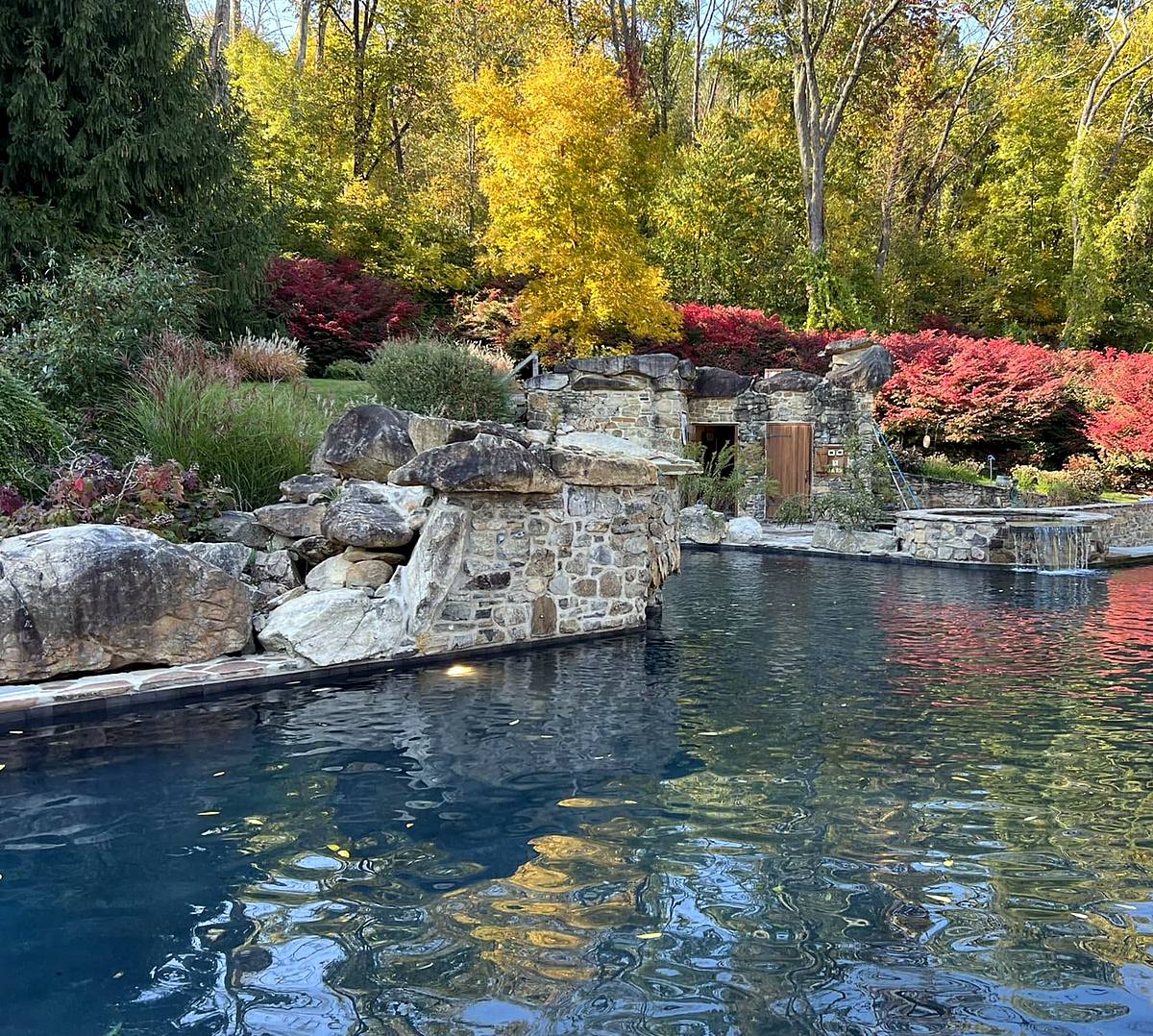 Natural boulder waterfall and stone spa spilling into a pool, with fall foliage behind