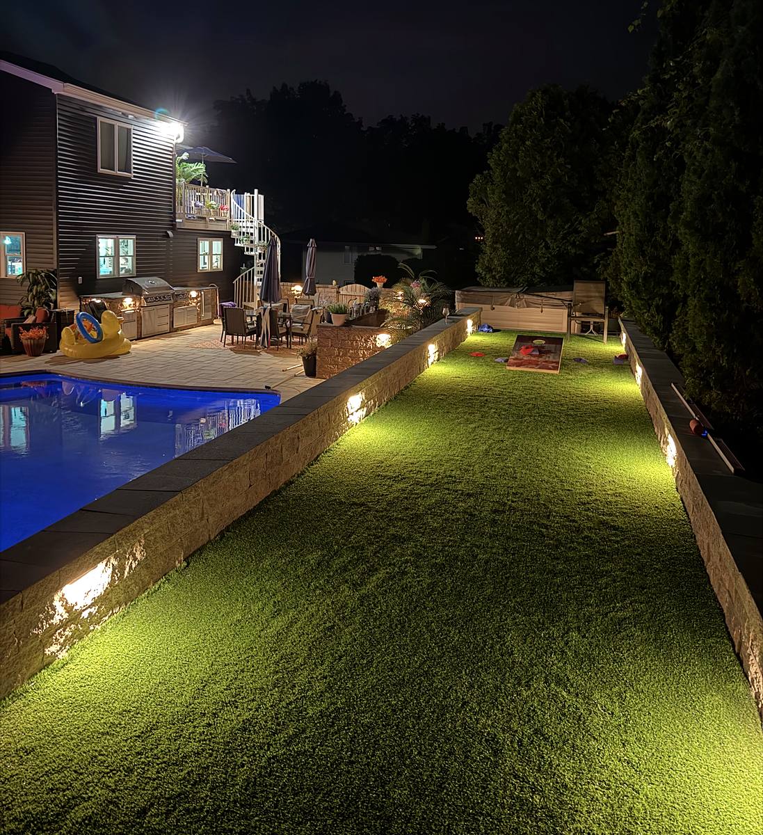 Backyard at night: a custom turf horseshoe pit between lit sitting walls, with the blue-lit pool and outdoor kitchen beyond